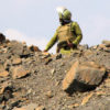 Tanzanian police officer in dull green riot gear and helmet, pictured against the sky guarding waste dump site at Barrick Gold's North Mara mine. Image courtesy Catherine Coumans / Mining Watch.